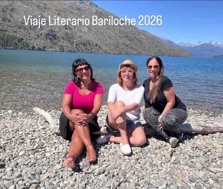Tres viajeras sentadas en la playa de piedras de un lago patagónico, con montañas nevadas detrás.