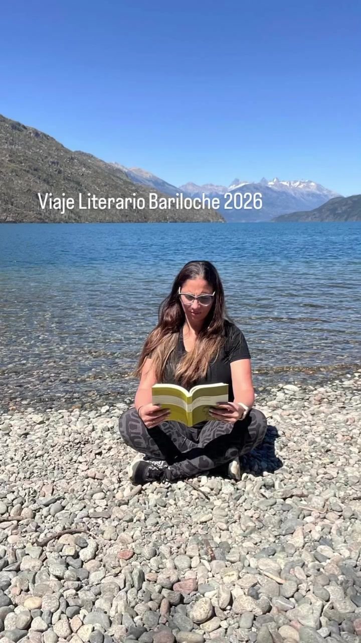 Una viajera lee sentada en la playa de piedras de un lago, con montañas de fondo.