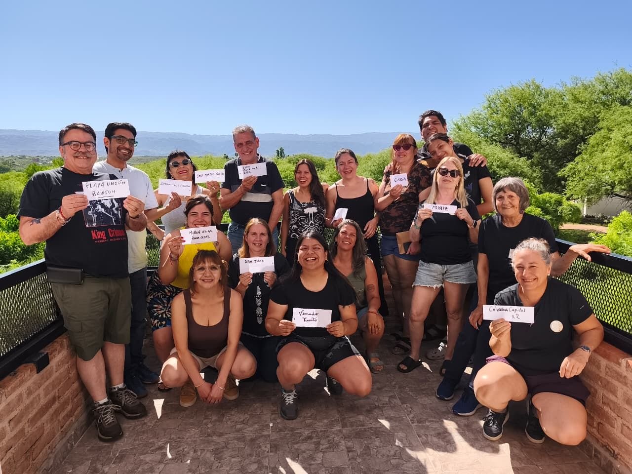 Grupo en un balcón con sierras de fondo; cada persona sostiene un cartel con el nombre de su ciudad.